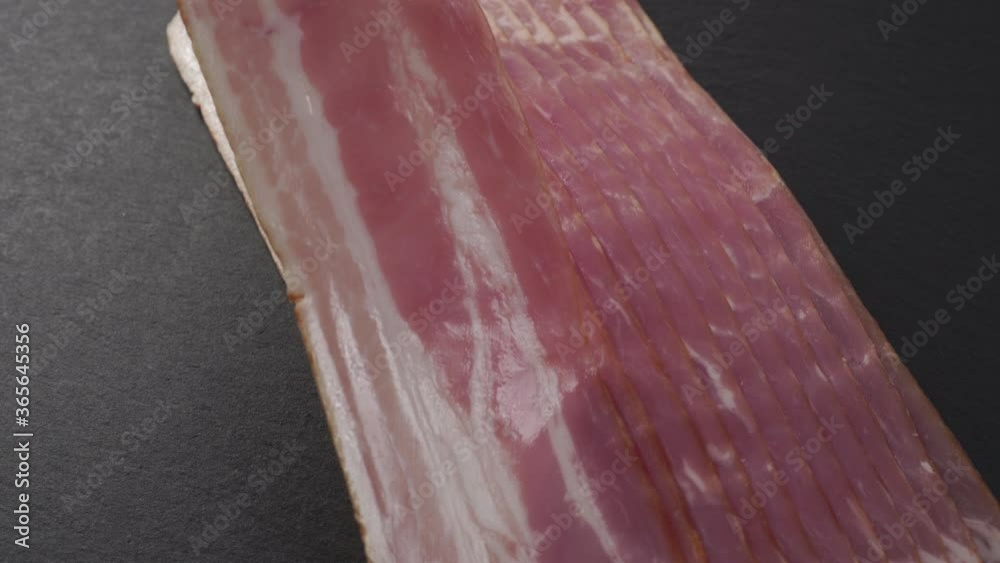 Hand takes a strip of fresh red bacon with fat layers. On a black stone board. Sliced appetizing meat