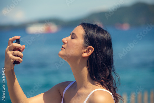 Young beautiful woman spraying thermal water or facial mist on her face at the beach. Summer skin care