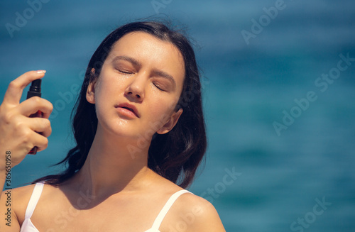Young beautiful woman spraying thermal water or facial mist on her face at the beach. Summer skin care