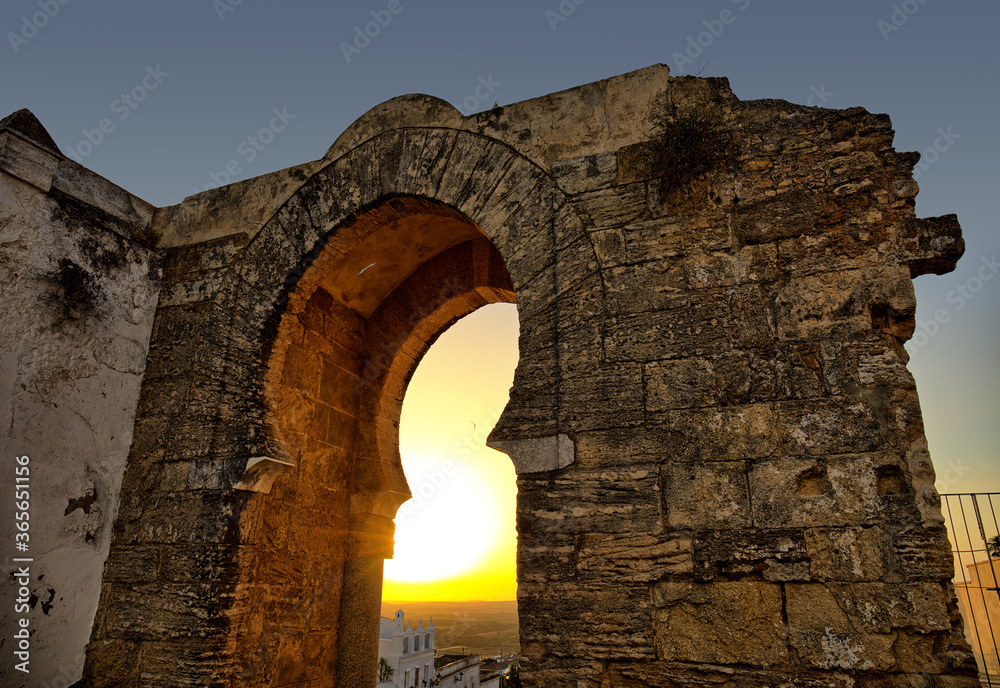 A romantic evening mood at the historic city gate of Medina Sidonia in Andalusia. The sun sets behind the stone gate.