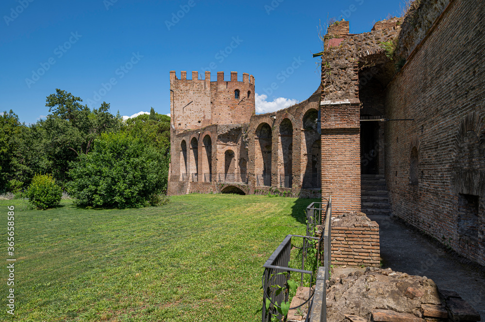 The Aurelian Walls of Rome at Porta San Sebastiano seen from inside the city. Brick support arches and cobblestone walkway. The walls border whit a vast lawn with trees. Clear day with clouds. Italy