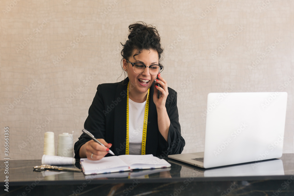 brunette woman with curly, upturned hair and round glasses smiling and talking on a mobile phone and writing on paper with a grey pen and a laptop next to her brunette woman with curly, upturned hair and round glasses smiling and talking on a mobile phone and writing on paper with a grey pen and a laptop next to her