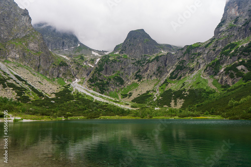 Beautiful view of foggy mountains cover by dark clouds and green forest with a reflection in a lake. Green mountain lake, Zelene Pleso, High Tatras, Slovakia