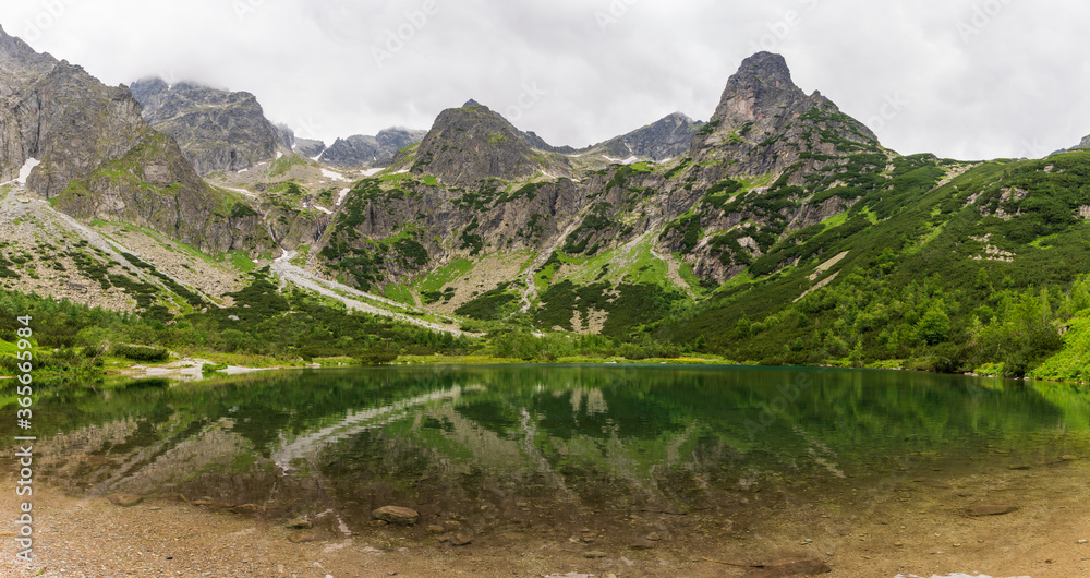 Naklejka premium Beautiful view of foggy mountains cover by dark clouds and green forest with a reflection in a lake. Green mountain lake, Zelene Pleso, High Tatras, Slovakia