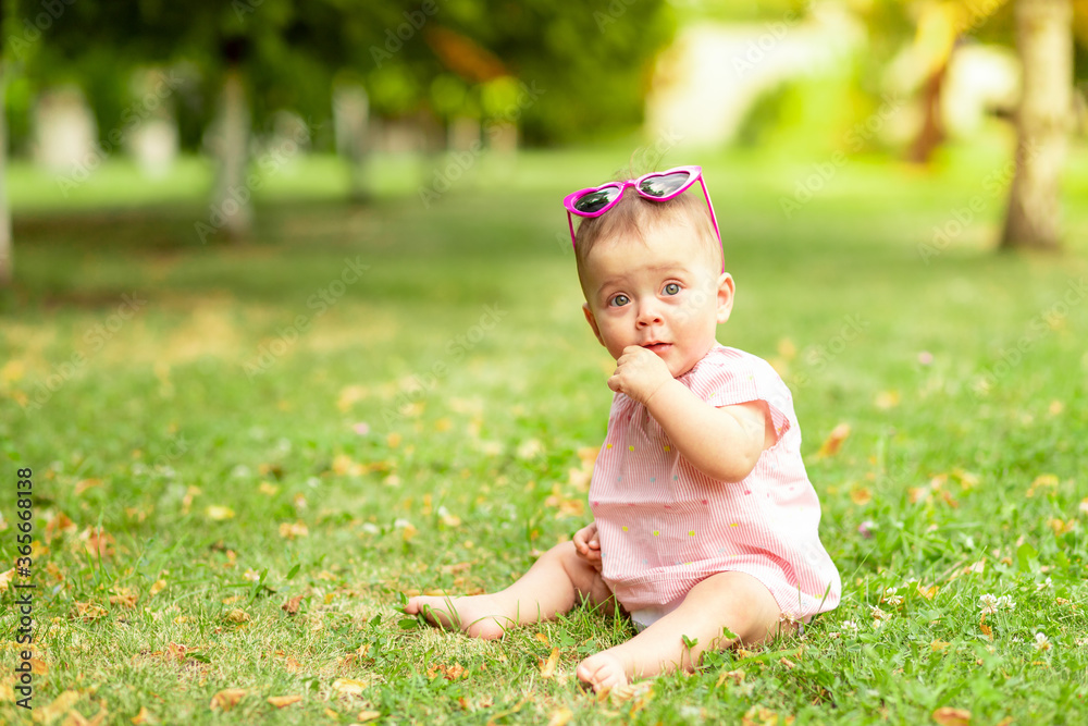 little baby girl 7 months old sitting on the green grass in a pink bodysuit and bright glasses, walking in the fresh air little baby girl 7 months old sitting on the green grass in a pink bodysuit and bright glasses, walking in the fresh air