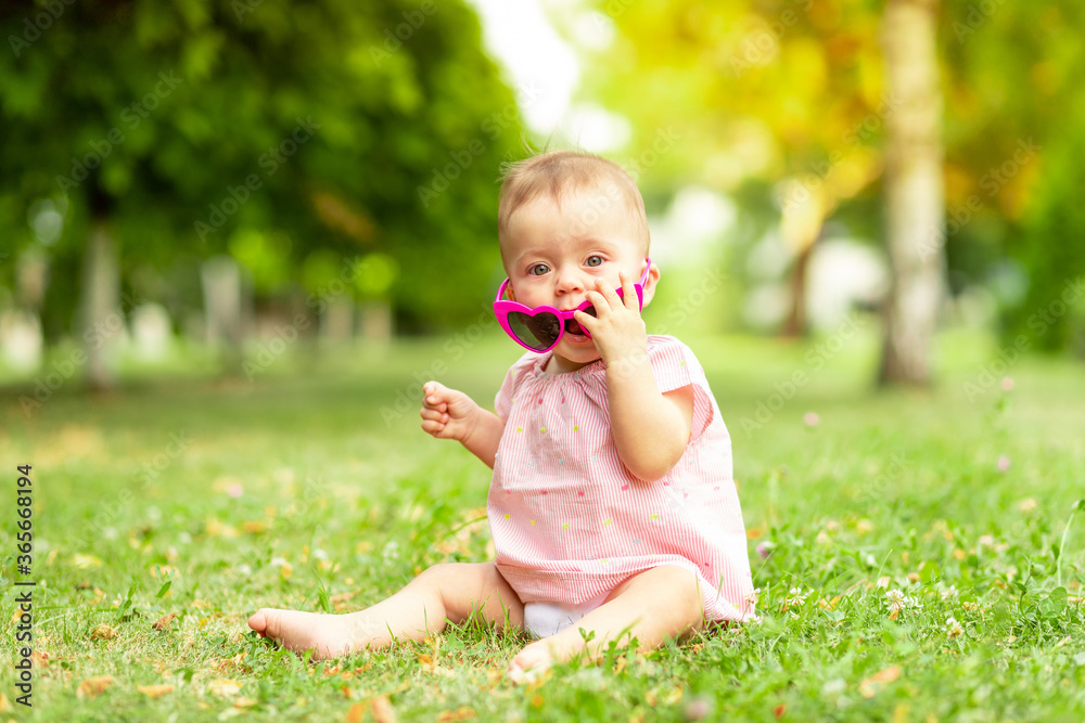 little baby girl 7 months old sitting on the green grass in a pink bodysuit and bright glasses, walking in the fresh air little baby girl 7 months old sitting on the green grass in a pink bodysuit and bright glasses, walking in the fresh air