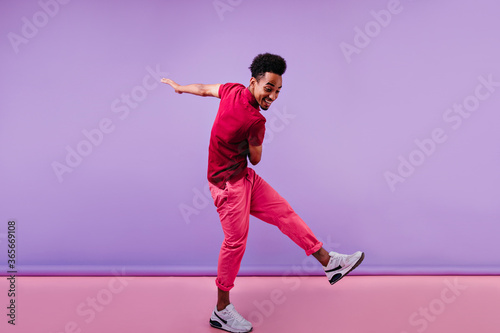 Full-length shot of trendy african male model in pink clothes. Excited black man in stylish sneakers dancing on purple background.