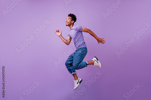 Portrait of jumping amazed guy in white sneakers. Indoor shot of dancing stylish male model in purple t-shirt. © Look!