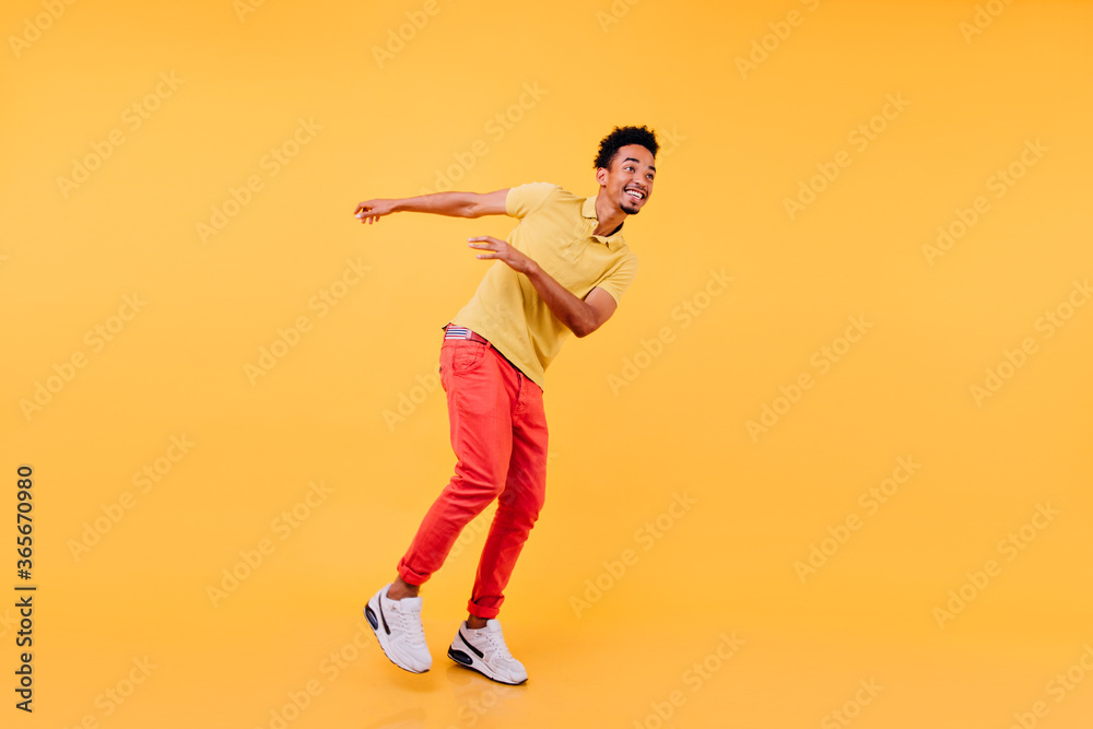 Pleased african man with sincere smile dancing on yellow background. Studio photo of enthusiastic black-haired guy wears white sneakers. Pleased african man with sincere smile dancing on yellow background. Studio photo of enthusiastic black-haired guy wears white sneakers.