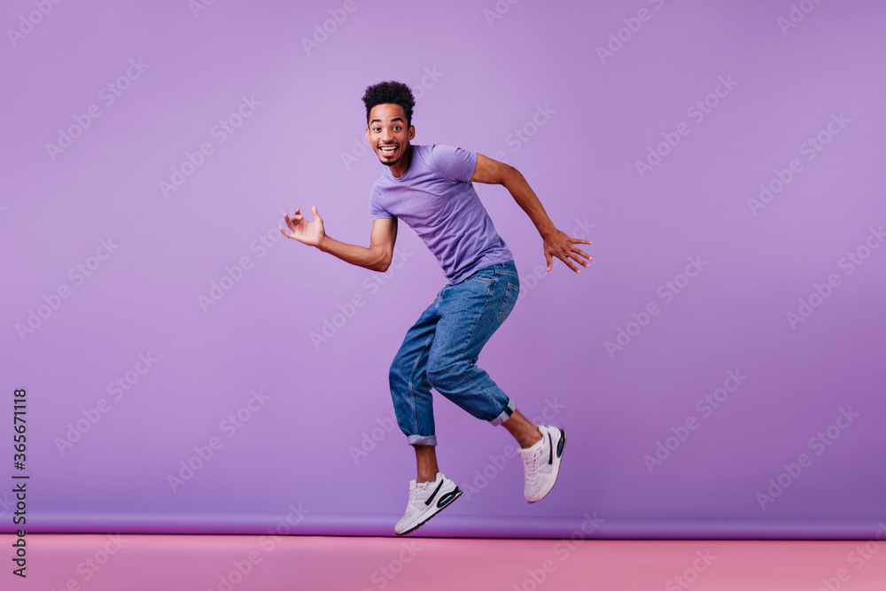 Good-humoured male model dancing in new white sneakers. Studio shot of ecstatic african guy in jeans.