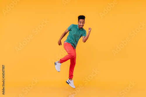 Full-length shot of inspired black guy dancing in red pants. Indoor photo of carefree african man enjoying photoshoot on yellow background. © Look!