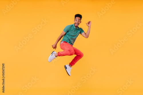 Joyful short-haired guy jumping on yellow background. Indoor photo of stunning male model in green t-shirt having fun in studio.