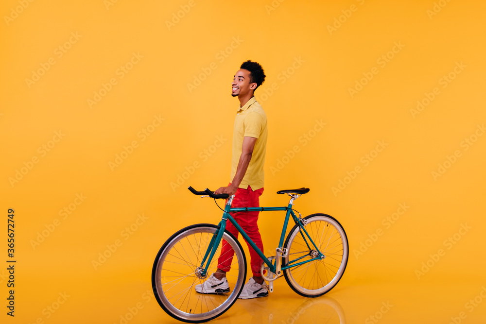 Full-length portrait of blithesome african man posing with bicycle. Photo of trendy black guy in sport shoes standing on yellow background. Full-length portrait of blithesome african man posing with bicycle. Photo of trendy black guy in sport shoes standing on yellow background.