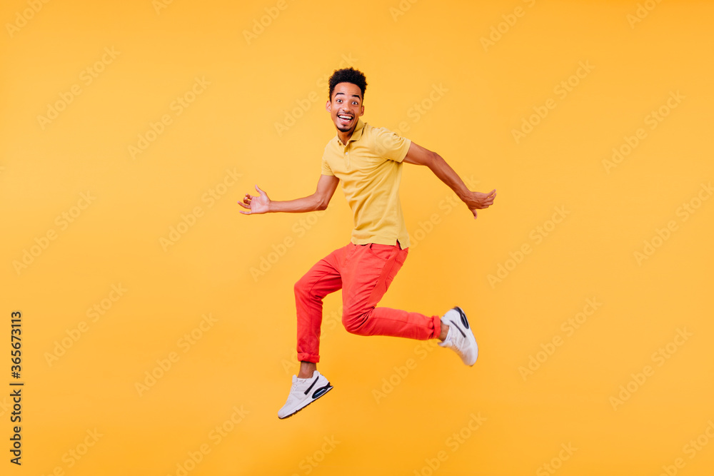 Dreamy male model with brown skin fooling around during photoshoot. Indoor shot of african guy in bright clothes jumping on yellow background.