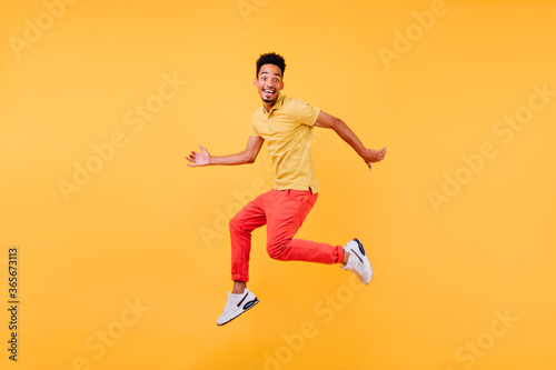 Dreamy male model with brown skin fooling around during photoshoot. Indoor shot of african guy in bright clothes jumping on yellow background.