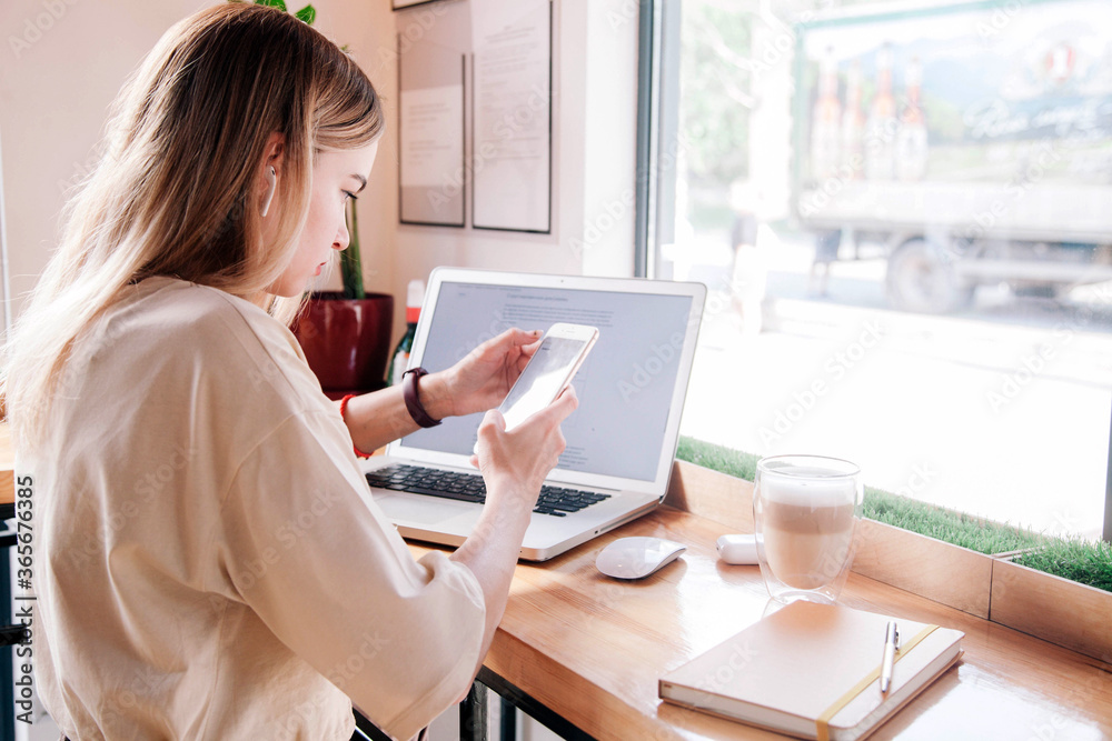 Young girl with phone in hand works behind a laptop in a coffee shop and drinks a cappuccino Young girl with phone in hand works behind a laptop in a coffee shop and drinks a cappuccino