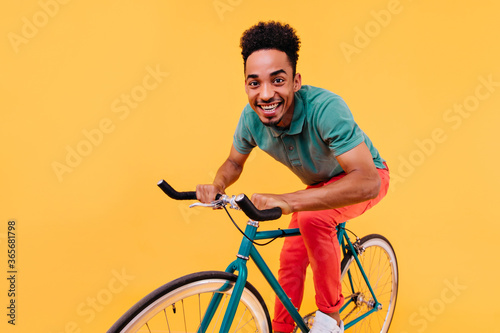 Active african guy in green t-shirt sitting on bike. Laughing black young man posing with bicycle on bright background.