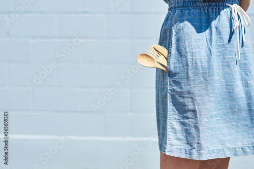 A women side pocket on a dress with reusable wooden, bamboo cutlery. On a bright sunny day. Healthy lifestyle and eco friendly, Reuse and recycle concept. Blue dress and brick wall background.