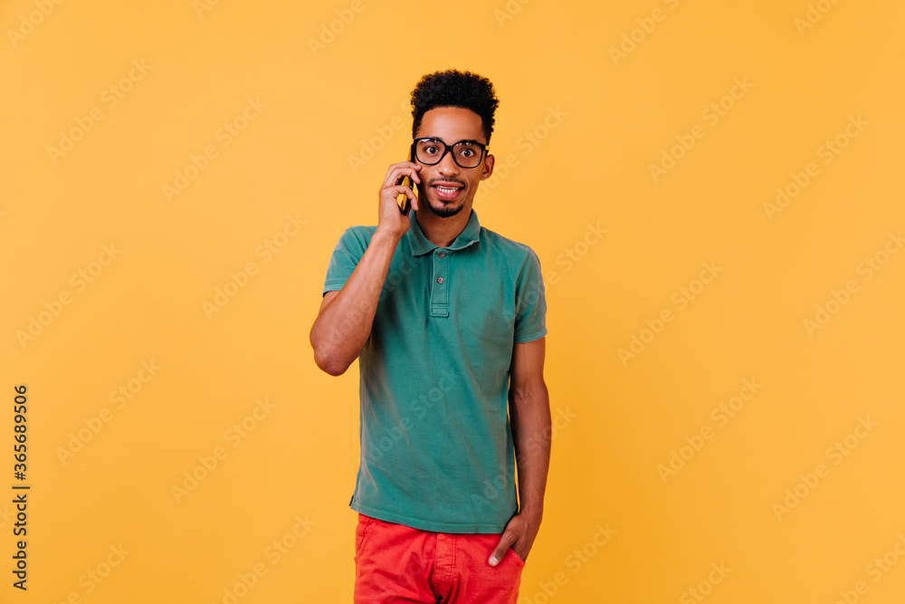 Surprised black male model talking on phone. Studio shot of stylish african guy in glasses posing on yellow background with smartphone. Surprised black male model talking on phone. Studio shot of stylish african guy in glasses posing on yellow background with smartphone.