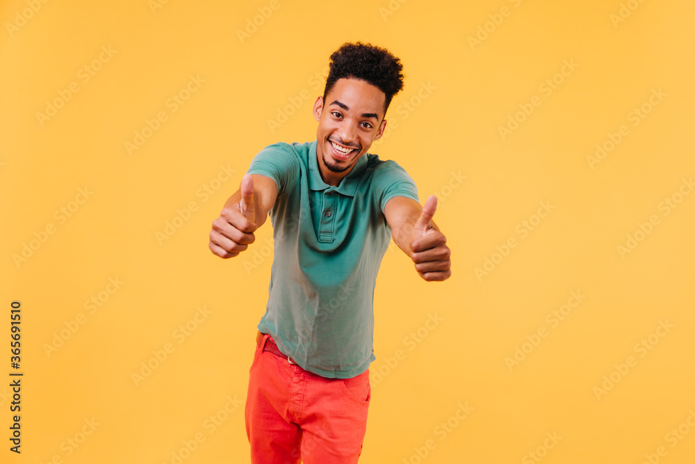 Funny short-haired man posing on yellow background with thumbs up. Indoor photo of happy african guy in trendy attire. Funny short-haired man posing on yellow background with thumbs up. Indoor photo of happy african guy in trendy attire.