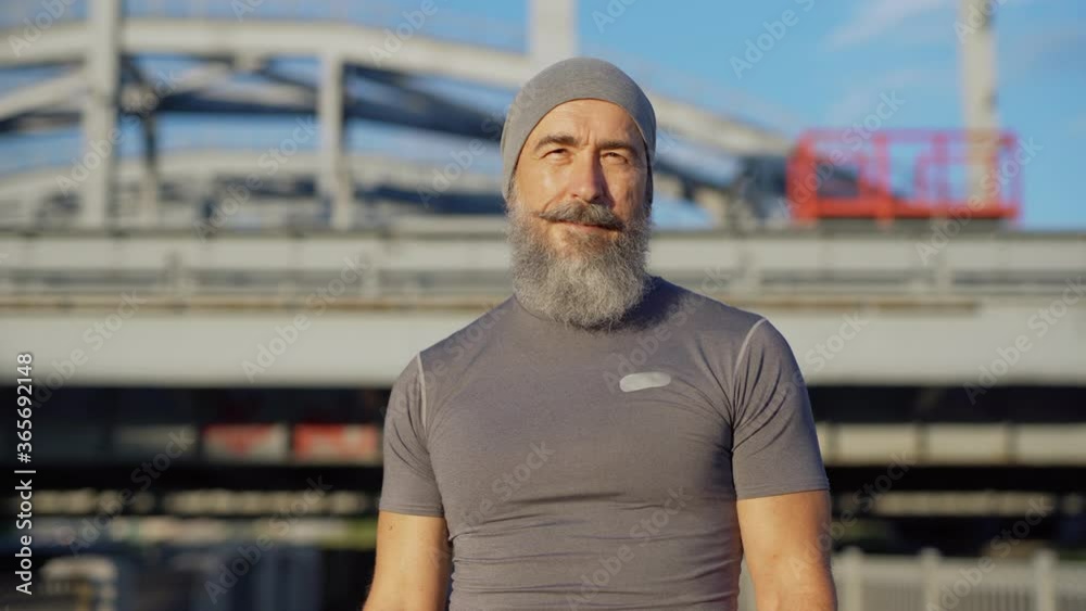 Tracking waist up portrait of senior athlete with grey beard looking at camera posing on bridge road after running workout Tracking waist up portrait of senior athlete with grey beard looking at camera posing on bridge road after running workout