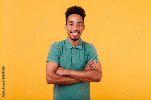 Laughing african guy with trendy haircut posing on bright yellow background with arms crossed. Studio shot of black man smiling to camera during photoshoot.