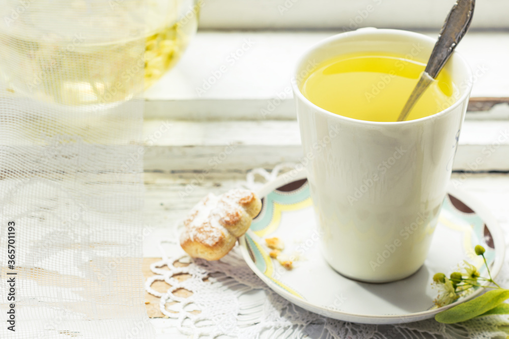 Linden flowers tea in a white cup with silver spoon on a white napkin, on a wooden background and cookie nearby, yellow drink of linden flower on a vintage background, healthcare and healthy eating co Linden flowers tea in a white cup with silver spoon on a white napkin, on a wooden background and cookie nearby, yellow drink of linden flower on a vintage background, healthcare and healthy eating co