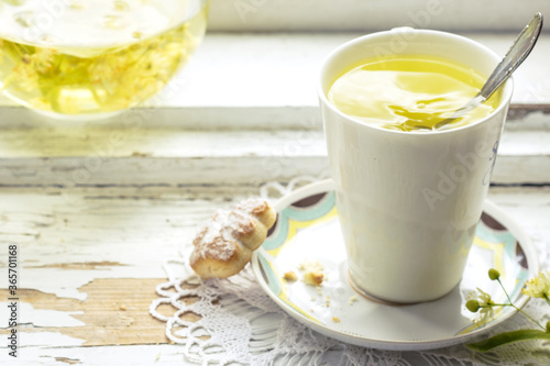 Linden flowers tea in a white cup with silver spoon on a white napkin, on a wooden background and cookie nearby, yellow drink of linden flower on a vintage background, healthcare and healthy eating co