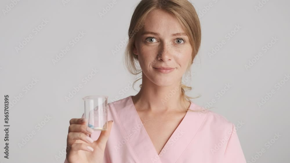Young beautiful nurse holding medical pills confidently looking in camera over gray background Young beautiful nurse holding medical pills confidently looking in camera over gray background