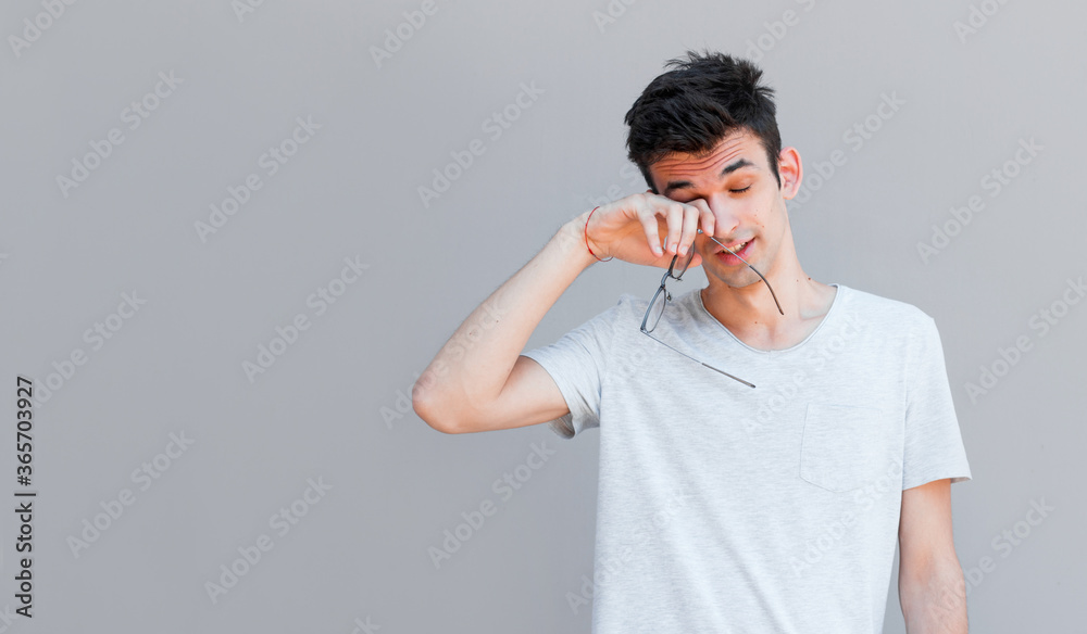 Portrait of tired ordinary european guy with bristle, holding glasses while rubbing eyes, being exhausted and wanting to get rest against gray background. Man spent too much time near laptop Portrait of tired ordinary european guy with bristle, holding glasses while rubbing eyes, being exhausted and wanting to get rest against gray background. Man spent too much time near laptop
