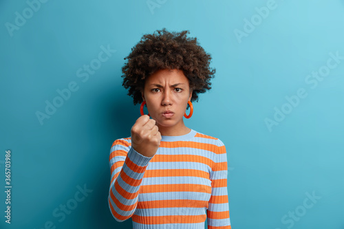 Disturbed angry woman clenches fist and has outraged expression, warns about revenge, expresses hate and negative emotions, wants punch enemy, dressed casually, isolated on blue studio wall.