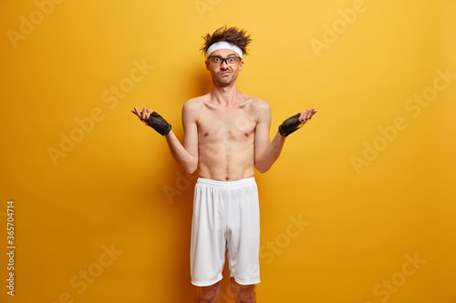 Puzzled hesitant man spreads hands and stands confused, wears white headband, sport gloves and white shorts, has workout or fitness training, poses with naked torso against yellow background