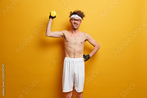 Body care and workout concept. Displeased European man holds sport equipment up, lifts dummbell with strength, dressed in shorts and gloves, puts efforts in gaining goal, leads active lifestyle photo