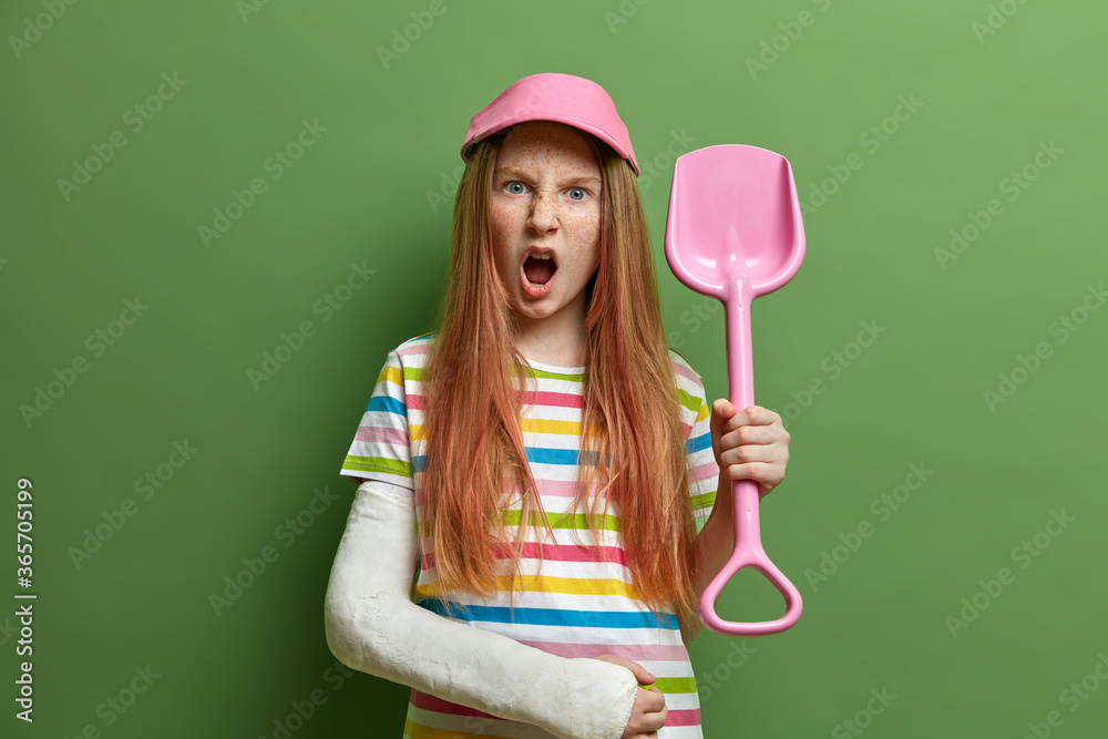 Φωτογραφία Angry small girl poses wit sand shovel, recreats at beach during summer time, has broken arm after accident, being irritated, disabled and displeased