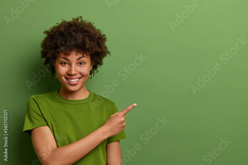 Friendly looking glad female shop assistant happy to help customers, shows way and demonstrates discounts in shop, points fore finger aside on empty space over green background. Advertising concept