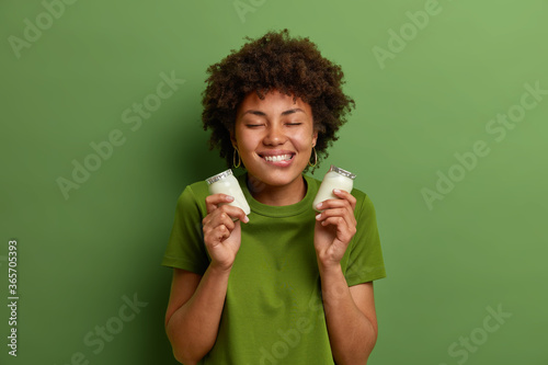 Portrait of cheerful woman holds two bottles of natural dairy yogurt for breakfast  bites lips and has temptation to eat  stands with closed eyes  isolated on green wall. Healthy nutrition concept