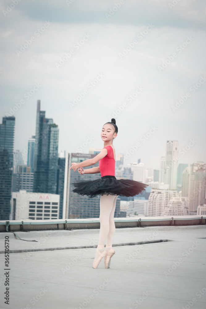Fototapeta premium Asian ballerina dancer girl practicing ballet dancing on rooftop with skyscraper city view, adorable child dancing in ballet
