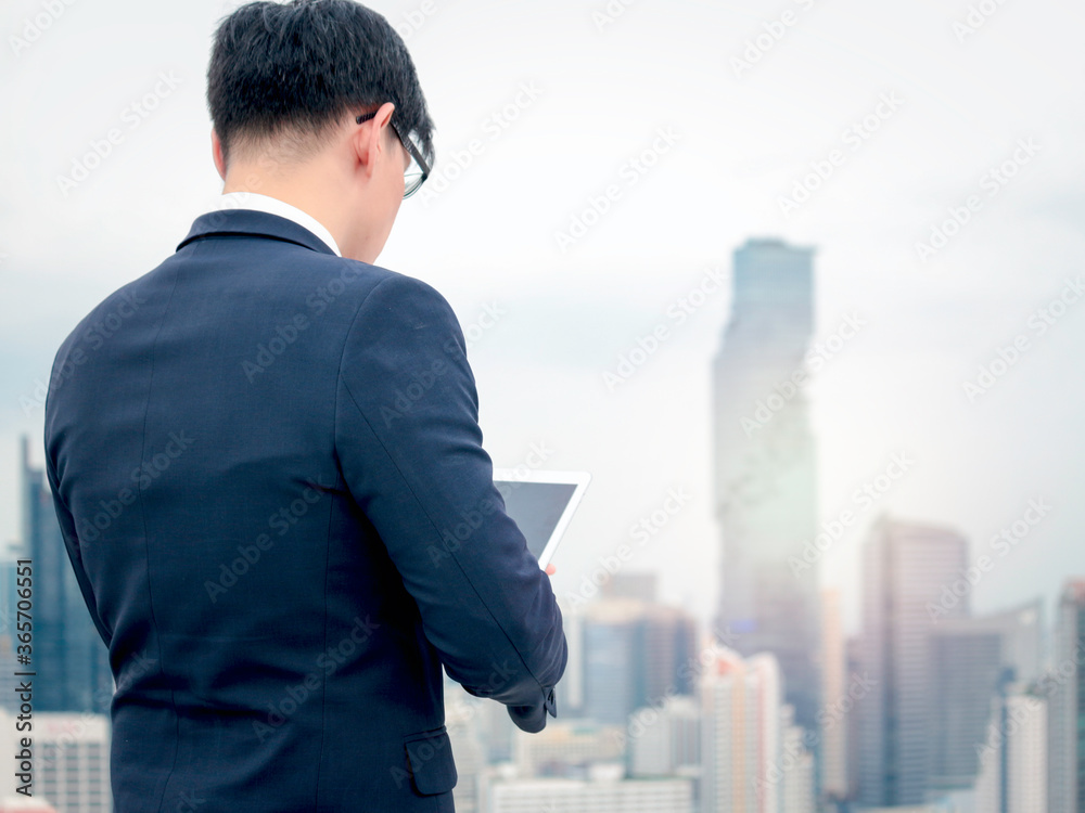 Young Asian businessman in suit using digital tablet on the rooftop with skyscraper city view
