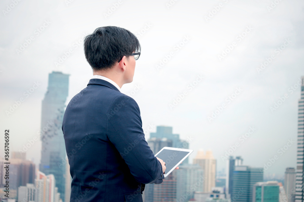 Young Asian businessman in suit using digital tablet on the rooftop with skyscraper city view Young Asian businessman in suit using digital tablet on the rooftop with skyscraper city view