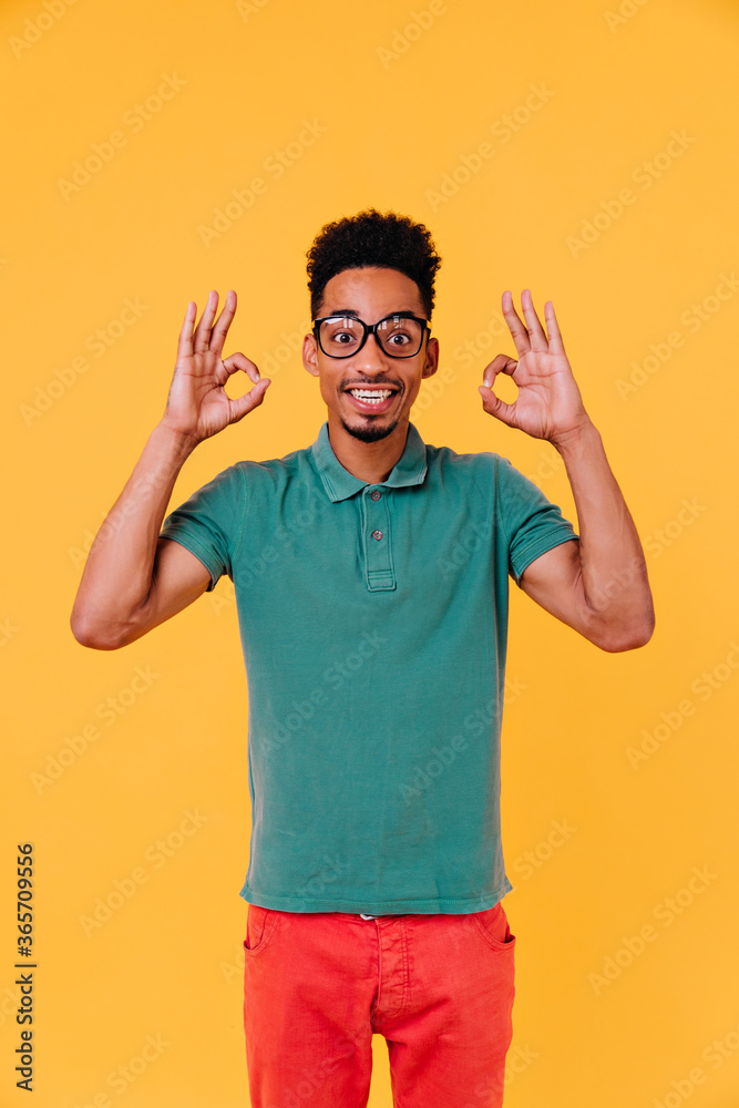 Naklejka premium Inspired young man posing with surprised smile and making okay sign. Studio portrait of enthusiastic guy in colorful attire and glasses.