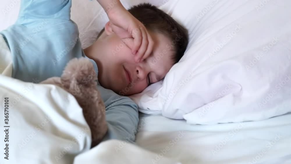 caucasian child boy in blue pajama hugging teddy bear wakes up from sleep in the morning. Kid yawning and stretching in a white bed after wake up. male toddler rubbing eyes and stretching in a bedroom caucasian child boy in blue pajama hugging teddy bear wakes up from sleep in the morning. Kid yawning and stretching in a white bed after wake up. male toddler rubbing eyes and stretching in a bedroom