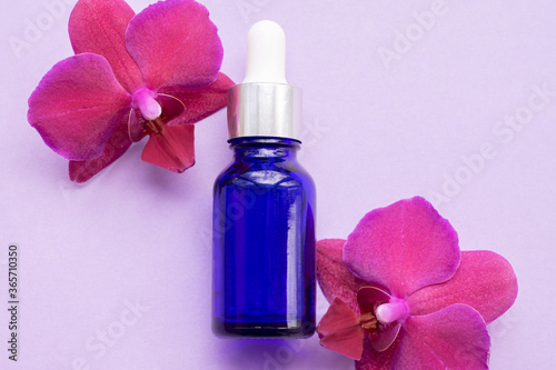 Blue glass bottle filled by essence or serum with orchid extract on purple background with bright blossoming orchids phalaenopsis. Eco cosmetic concept