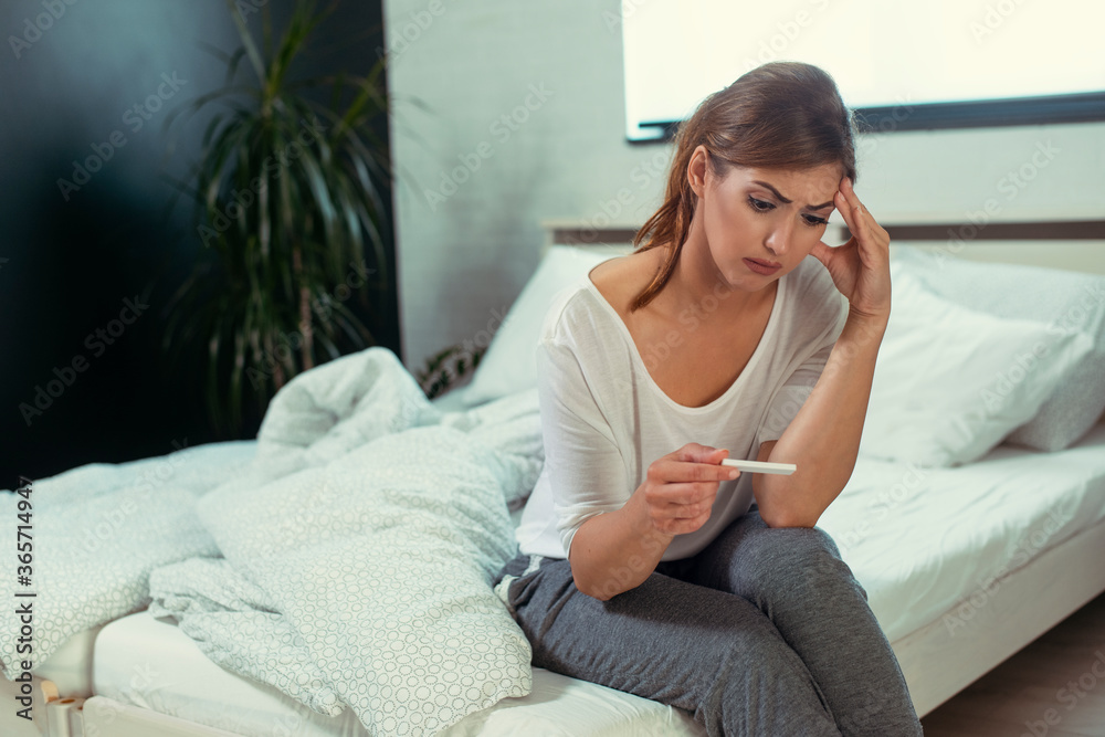 Upset woman sitting sad in white bed with negative pregnancy test