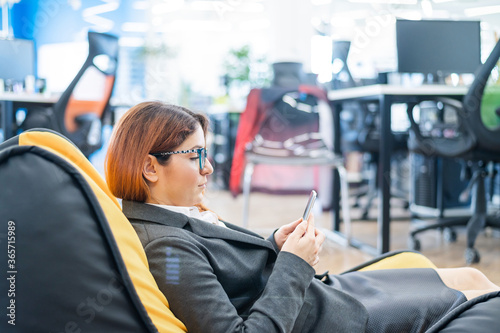 Business woman resting in a cozy bag chair and enjoys mobile. Female Manager at a break in a modern office. Caring for the convenience of employees. Beanbag in the workplace.