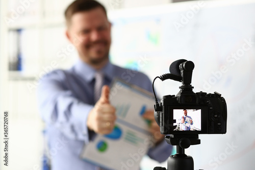 Male in suit and tie show confirm sign arm making promo videoblog or photo session in office camcorder to tripod portrait. Vlogger promotion selfie solution or finance advisor management information