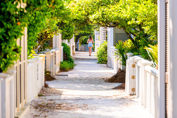 Seaside, Florida white wooden beach architecture path way with green landscaped shrubs bushes in sunlight and young woman in background distance