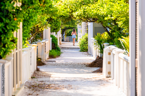 Seaside, Florida white wooden beach architecture path way with green landscaped shrubs bushes in sunlight and young woman in background distance