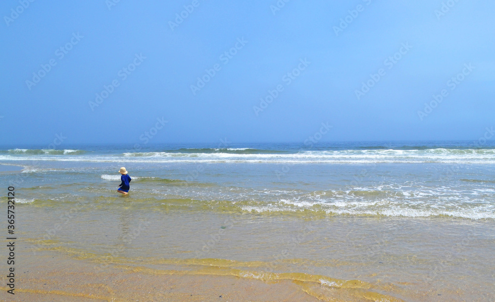 Ocean beach landscape against blue sky with small child  playing at water's edge in small waves wearing bathing suit and hat. Summer blue sky with copy space.