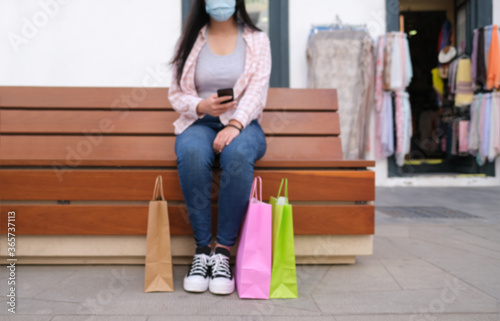 Blurred woman wearing face mask and using mobile phone - Asian girl sitting on a bench after shopping - Coronavirus lifestyle - Defocused image photo