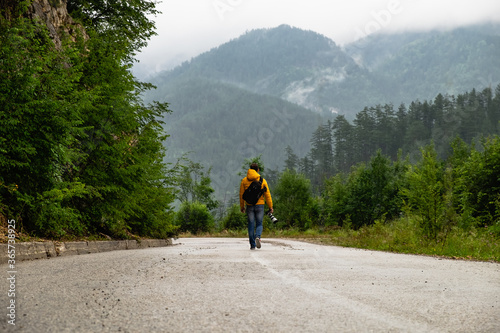 A man in yellow jacket and jeans with backpack and camera is walking alone down the road in the misty mountains in the distance and trees surrounding the road and covering mountain slopes